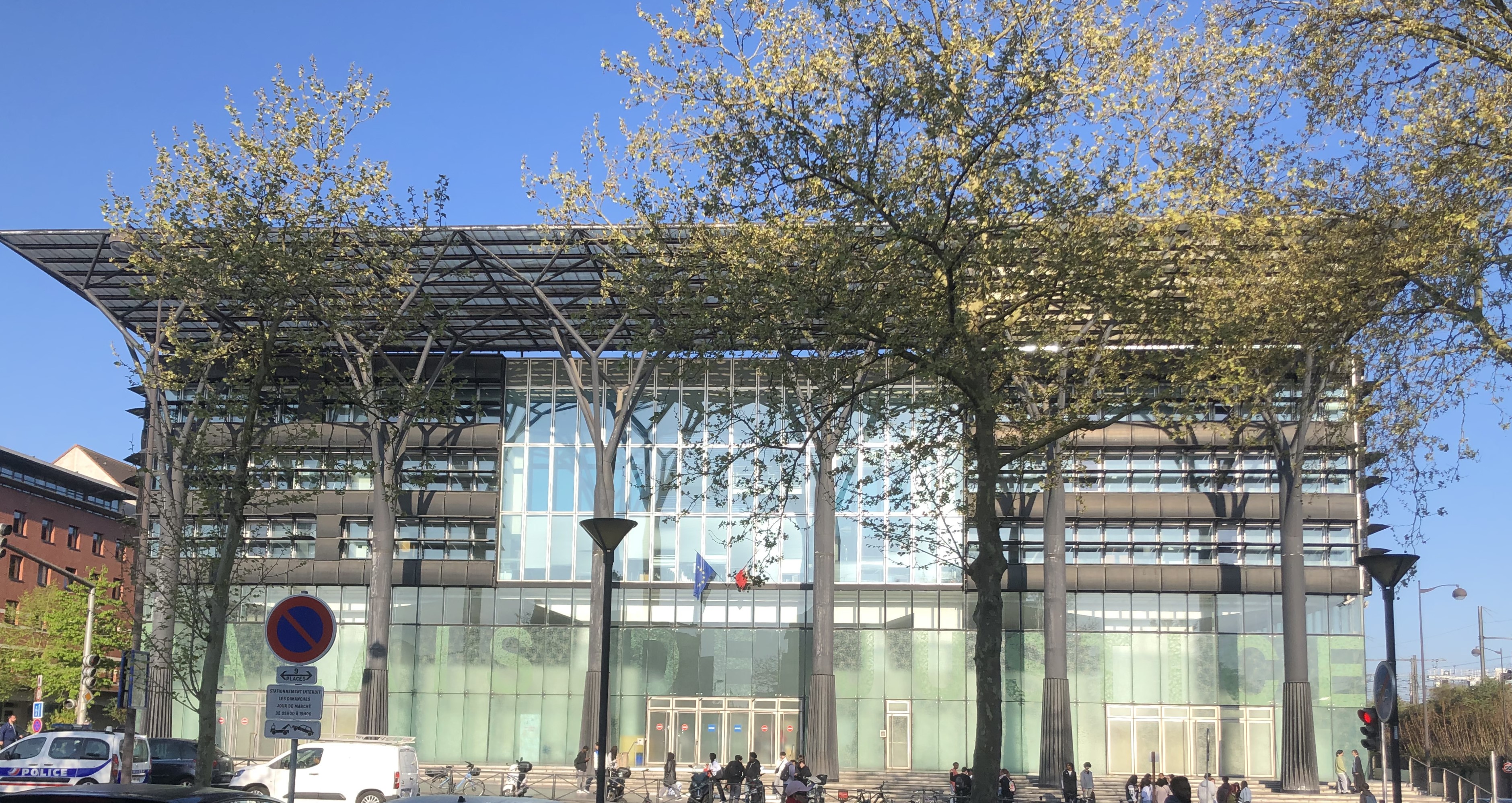 Vue extérieure du tribunal de Melun. Un bâtiment moderne et vitré, reconnaissable à son imposant auvent en métal et ses colonnes noires, vu depuis la rue, à travers les arbres de l'avenue.
