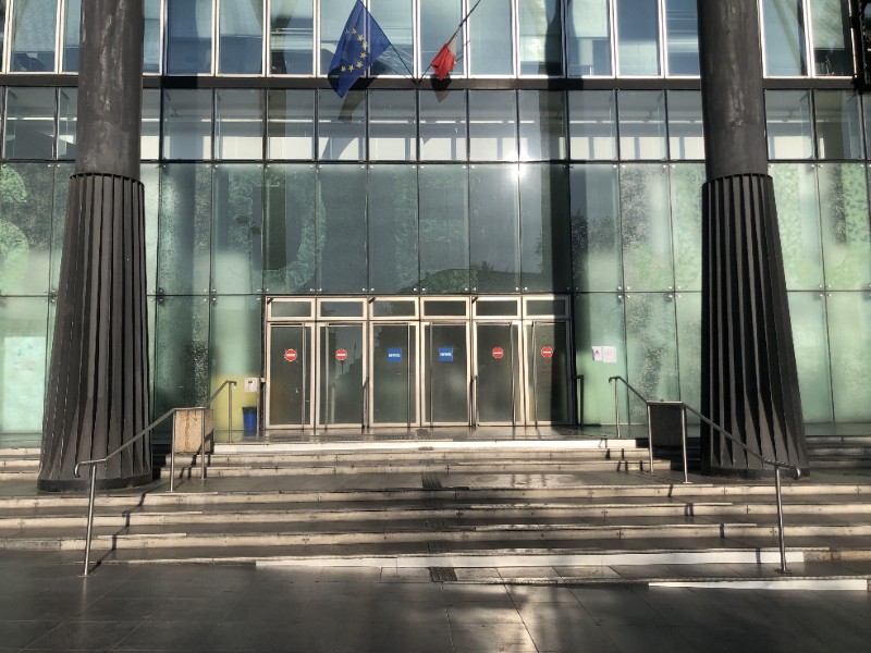 Entrée du tribunal avec ses portes vitrées encadrées par les drapeaux français et européen, en haut de quelques marches d'escalier.