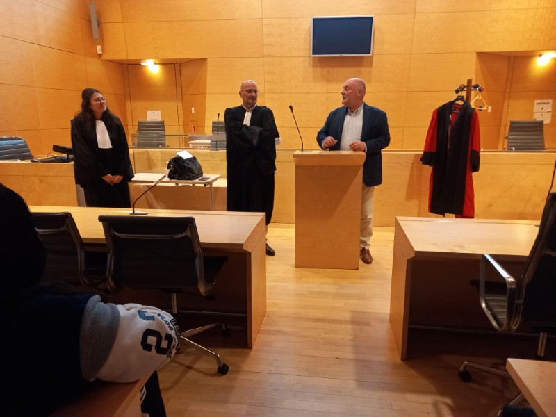 Trois magistrats en tenue, dont l'un en civil, se tiennent face à l'assemblée. La robe rouge est bien visible à côté du pupitre.