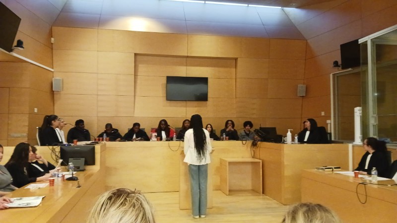 Une élève, se tient debout, de sos derrière un pupitre pour répondre aux questions de la cour. Face à elle, une douzaine de camarades en robes d'audience sont assis sur les bancs du tribunal, dans la salle d'audience boisée.