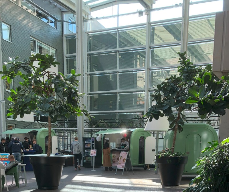 L'image présente des food trucks installés à l'extérieur du Deltion College, utilisés pour les pratiques commerciales et culinaires des étudiants. Ces camions sont aménagés pour la vente de nourriture, et l'espace autour d'eux est bien aménagé avec des tables et des chaises.