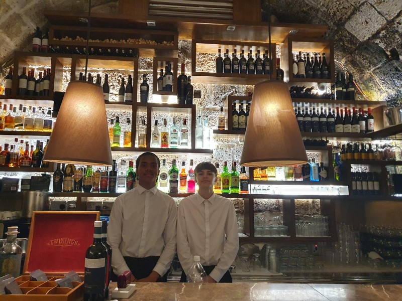 Deux élèves en tenue de service derrière un comptoir de bar garni de bouteilles.