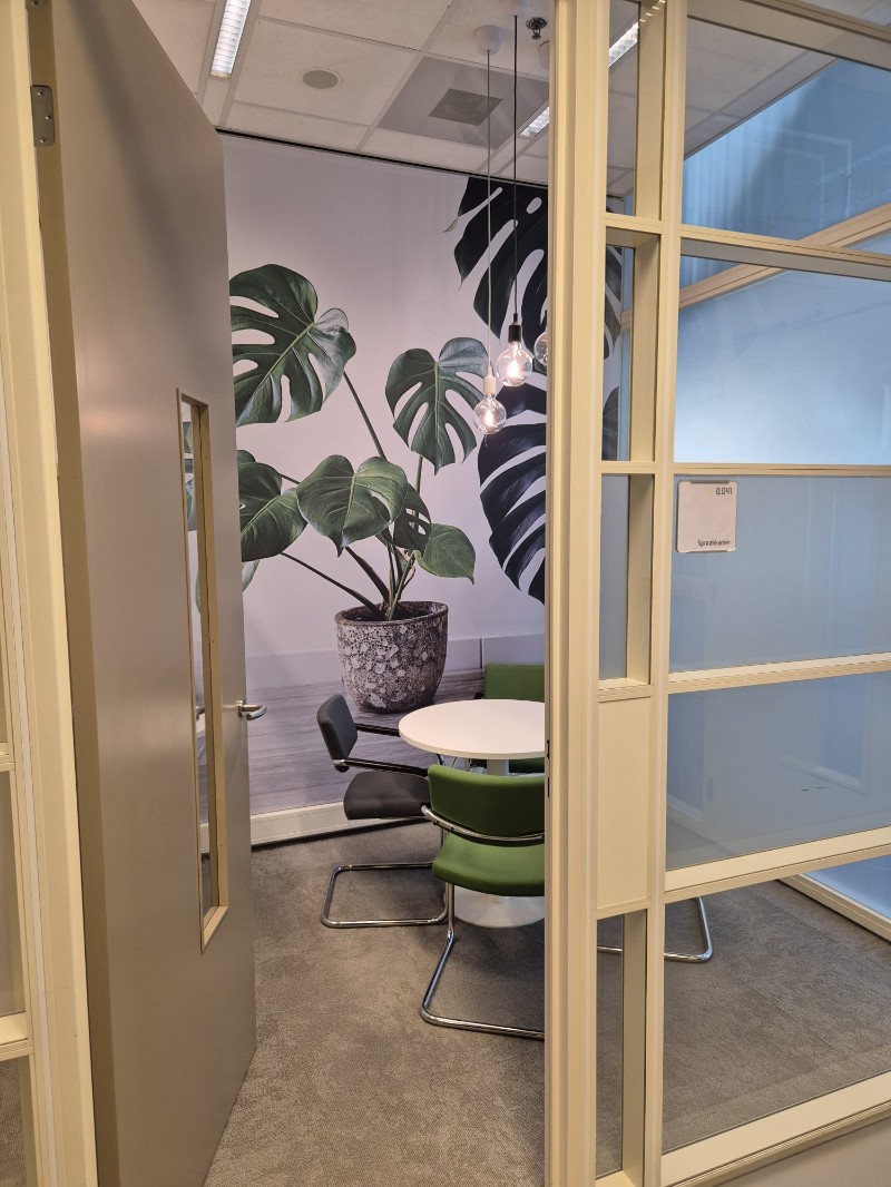 Photo d'un petit bureau moderne avec un mur en verre transparent avec la porte est partiellement ouverte, on voit une table ronde blanche avec des chaises vertes autour, dans un espace lumineux et épuré.