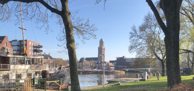 Cette photo présente un paysage paisible le long d'un canal à Zwolle. Des arbres bordent le canal. Le ciel est dégagé et la lumière naturelle.