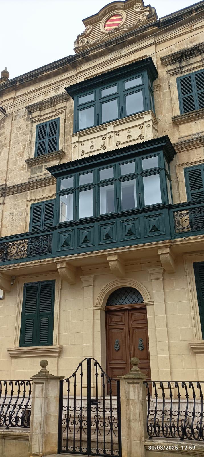 Façade en pierre avec volets verts foncés et un grand balcon fermé ; grille en fer forgé devant la porte.