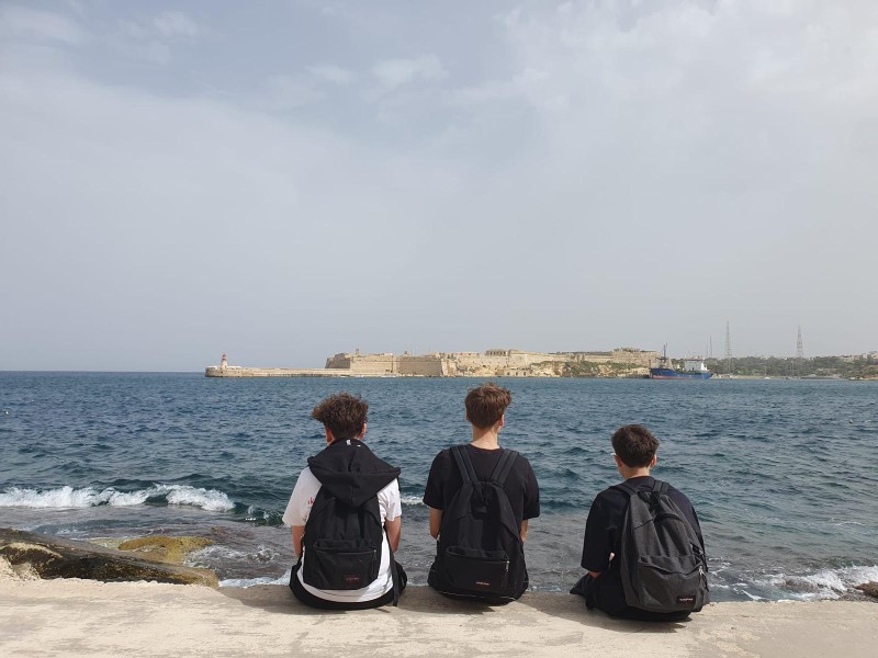 Trois élèves assis face à la mer, dos tourné, regardant une fortification au loin.