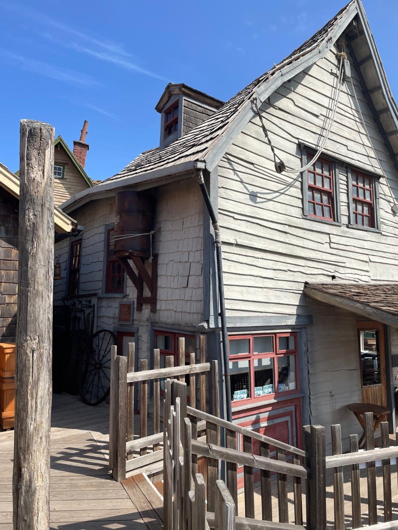 Maison en bois gris clair au toit pentu dans le Popeye Village, avec passerelles en bois au premier plan.