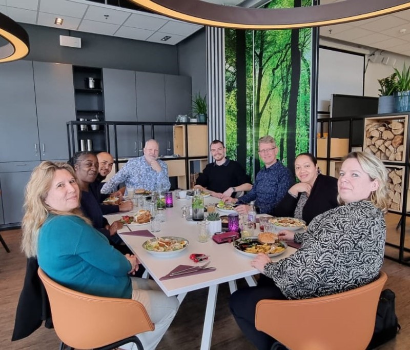 Cette image montre un repas pris par l'équipe franco-néerlandaise dans le restaurant d'application du Deltion College. L'équipe, composée de huit personnes (4 femmes e 4 hommes), est réunie autour d'une table pour un déjeuner-réunion, dans un cadre chaleureux et convivial.