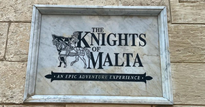 Plaque gravée fixée sur un mur en pierre indiquant "The Knights of Malta – An Epic Adventure Experience", devant un site touristique dédié à l'histoire de l'ordre des chevaliers de Malte.