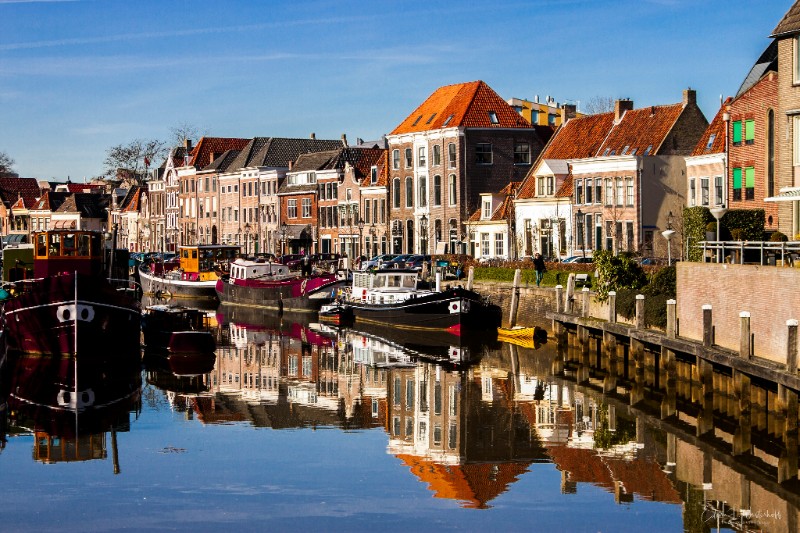 L'image présente une vue d'un canal traversant la ville, avec des bateaux amarrés sur les rives. On peut observer des bâtiments aux façades traditionnelles néerlandaises, avec des toits en tuiles rouges caractéristiques.
