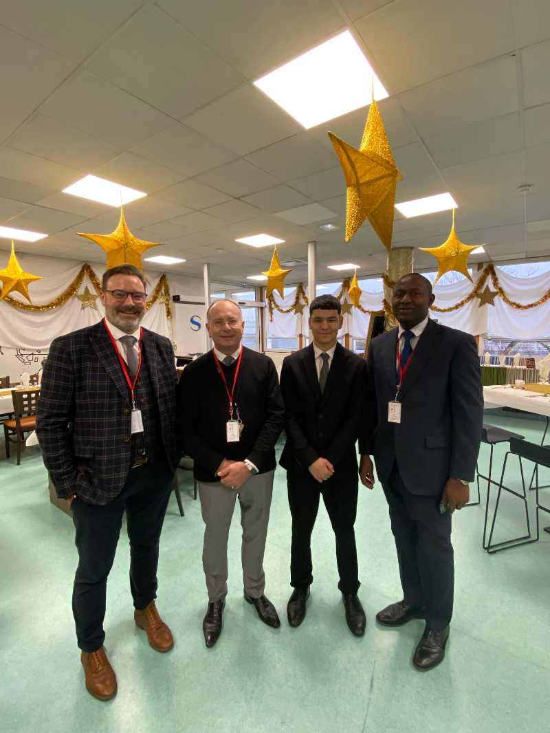 Mostafa Mohammadi en costume, souriant et posé entre trois enseignants de cuisine et de service, lors du TP organisé pour la SNECMA. La photo est prise dans un décor de brasserie décorée pour un événement festif.